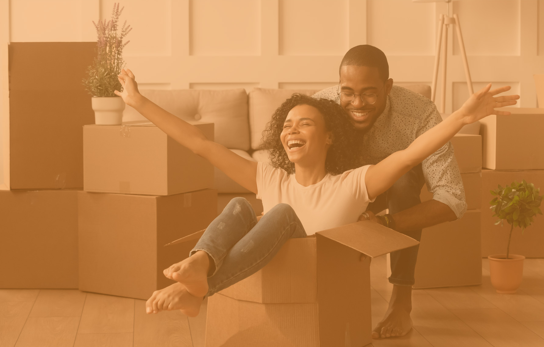 Happy african american young couple first time home buyers having fun unpacking laughing on moving day, excited wife riding sitting in cardboard box while black husband push it in new house apartment